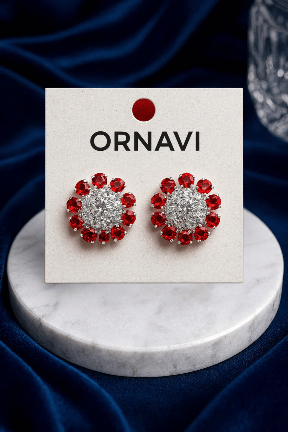 Heart-shaped earrings with red and clear stones on an ORNAVI branded card, set against a dark blue fabric background.