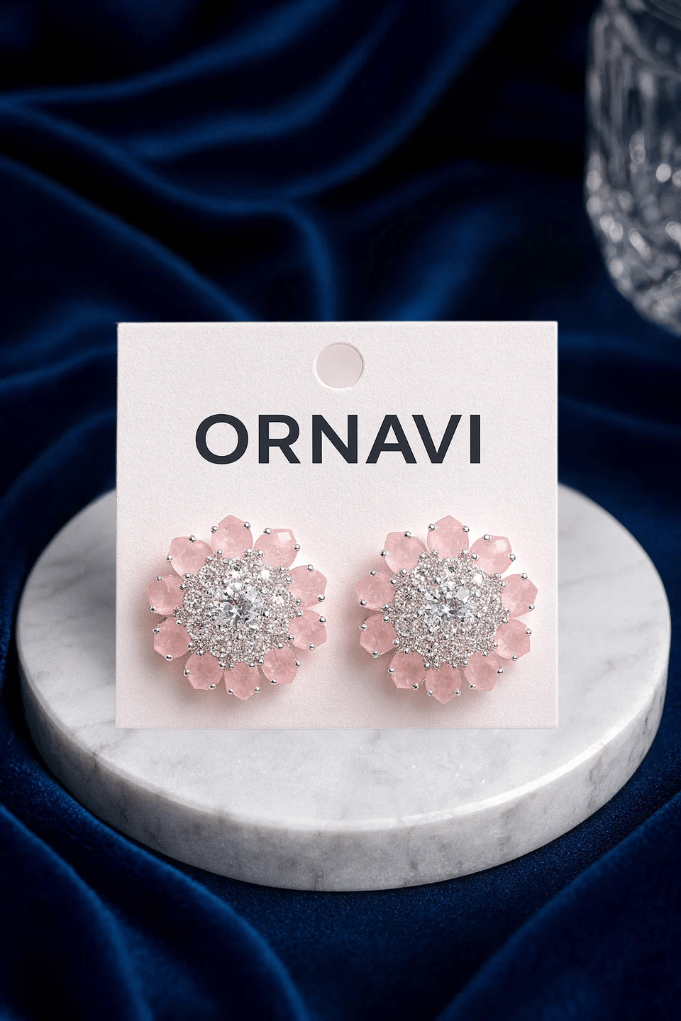Ornavi earrings with pink floral design on a marble stand against a dark blue fabric background