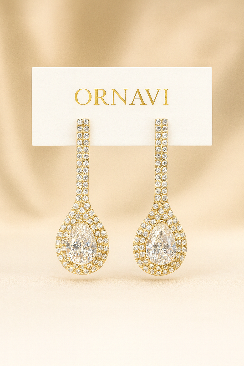 Pair of gold earrings with clear gemstones on a beige background with 'ORNAVI' card.