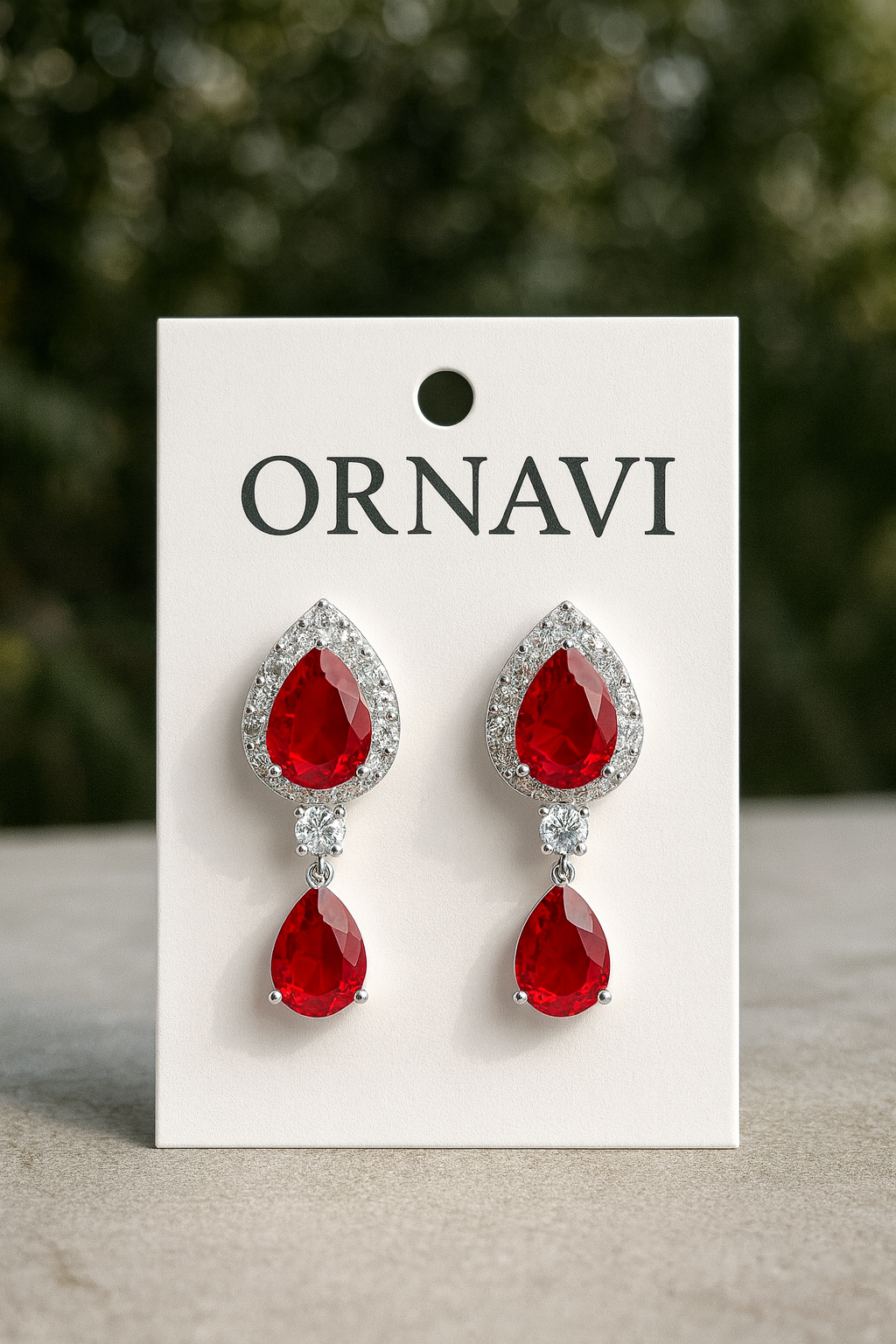 Red teardrop earrings on a white ORNAVI branded card with a blurred green background