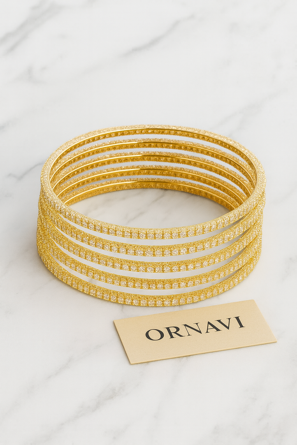 Set of gold bangles on a white marble surface with 'ORNAVI' card.